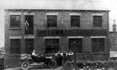 Black and white photo of the outside of Wm. Lamb Clog Manfuacturer, a rectangular brick building, two floors high, with large windows. There is a clogger stood at the first floor opening, a mustachioed man in a car loaded presumably with clogs, and three other workers in flat caps stood outside posing for the camera