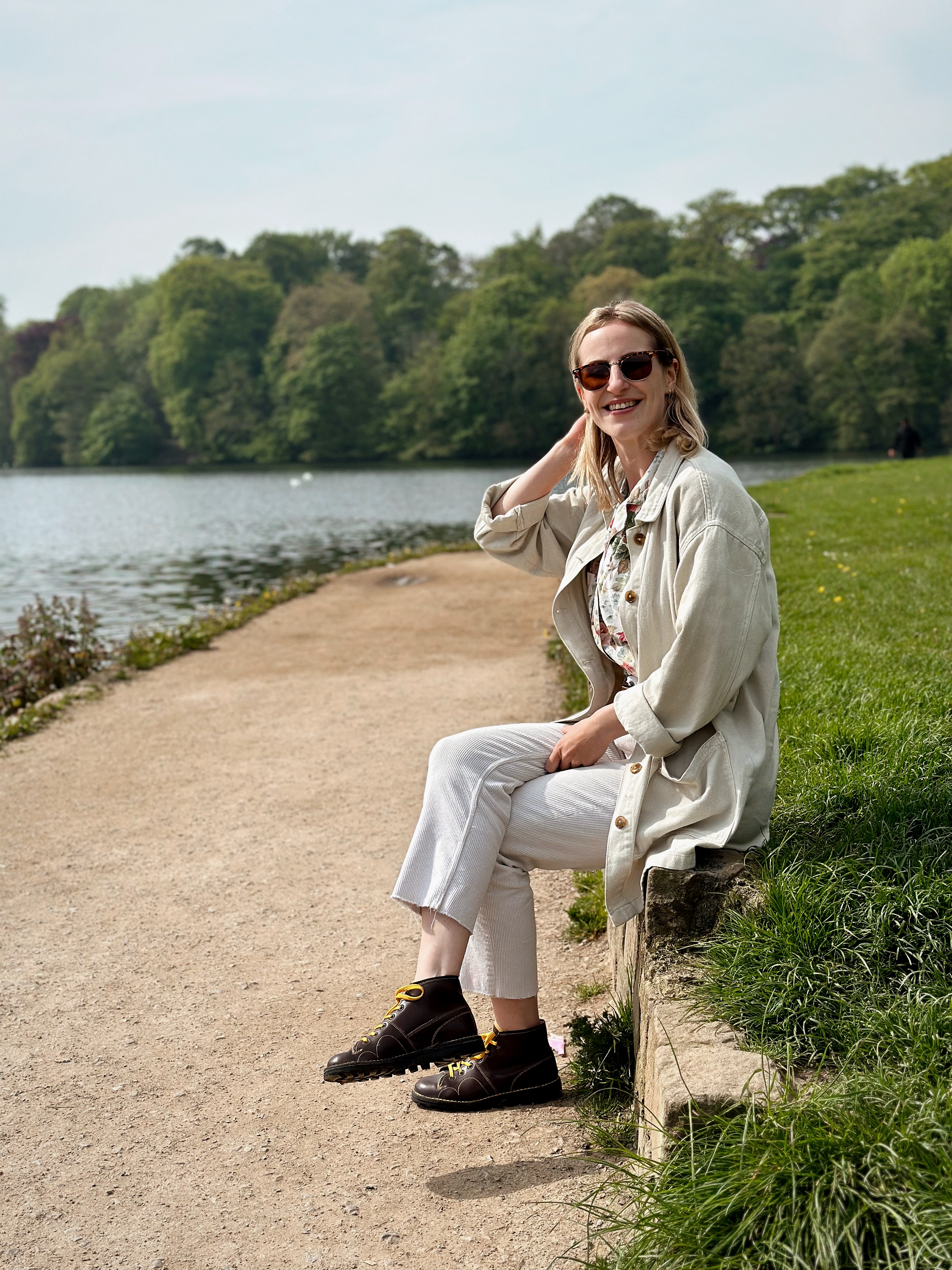 Amy is sitting on a low wall in front of a lake smiling to camera with her hand up to her hair. She is wearing cream trousers and a cream oversized jacket