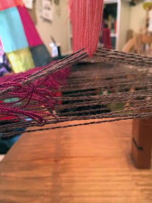 A view through the triangular tunnel formed by raised and flat grey warp threads. The raised threads are held up by salmon-pink loops of fine smooth string, and saturated fuchsia loops of the same string rest on the threads forming the left slope of the tunnel. The shadows make a complex, mostly-straight-line pattern on the wooden base of the loom.