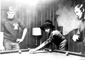 A photograph of Judy Garland, well-dressed and leaning over a pool table. There are two young men next to her.