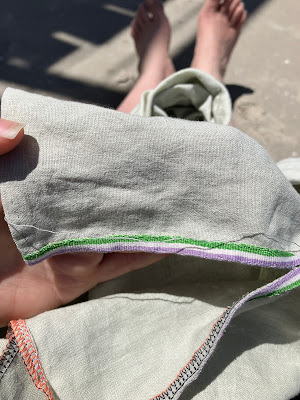 An in-progress picture of a woven ribbon being sewn in place on the side vent of a light green shirt. The ribbon is in the genderqueer pride flag colours of purple, white and green. Black and peach overlocking can be seen around the edges of the shirt pieces. End ID.