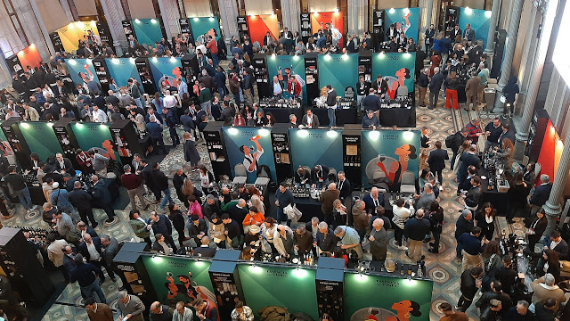 uma grande sala de evento com vários expositores de vinhos e muitas pessoas a circular e a provar vinhos