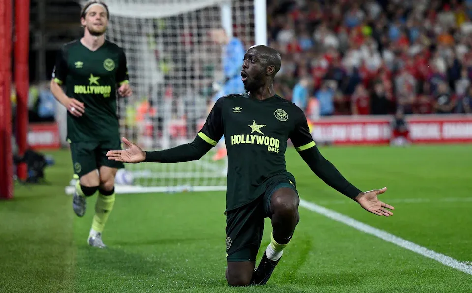 Jogador de futebol comemorando após marcar um gol, usando uniforme verde do Brentford. O estádio está lotado, destacando a empolgação da torcida