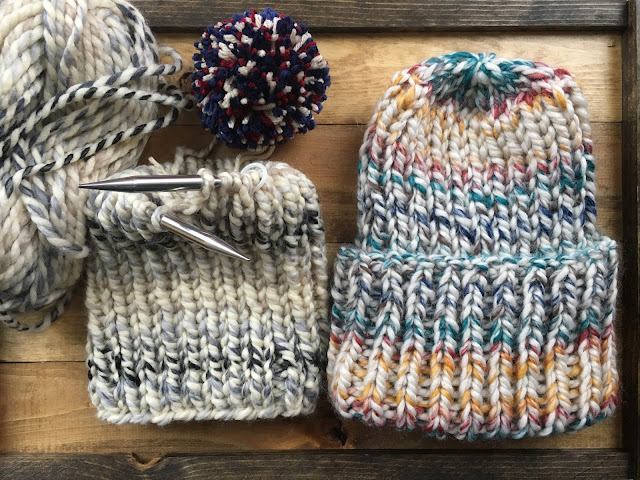 Two super bulky knit beanies laying on a wooden tray next to each other. Right one has teal, yellow, red, tan and folded up brim. Left one is still on circular knitting needles and are neutral shades. A large pom pom of navy, red, and tan awaits it fastening.