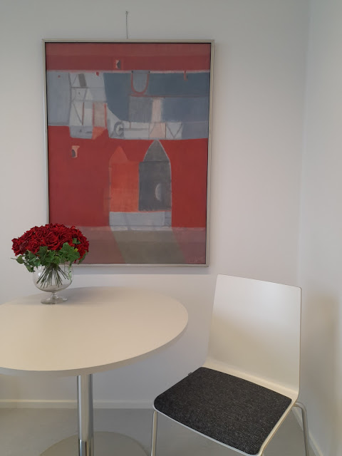 Kuvassa on tuoli, pyöreä pöytä, jossa on ruukussa oleva punainen kukkakimppu sekä seinällä pääasiallisesti punaisen värinen taulu.