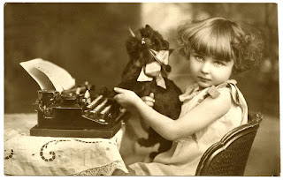 Vintage black and white photograph of a little girl sitting at a typewriter with a cat plushie.  The girl has the cat positioned so it looks as though the cat is the one typing.  