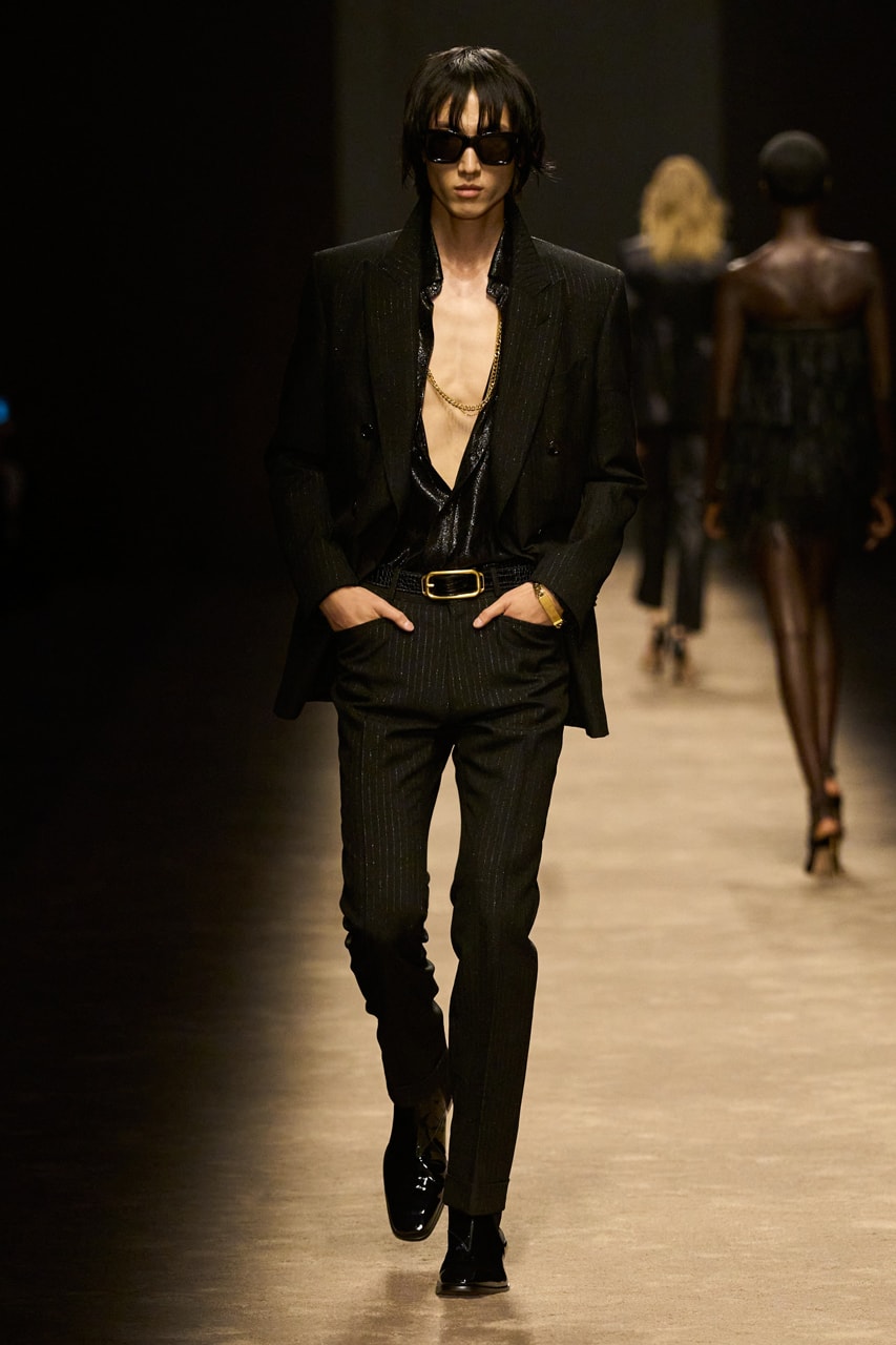 Man with black straight hair wearing black sunglasses, a gold chain necklace, a chunky gold id bracelet, a shiny black shirt unbuttoned, a black pinstripe blazer and trousers, and black patent leather laceup shoes walking on a taupe carpeted runway with his hands in his pockets
