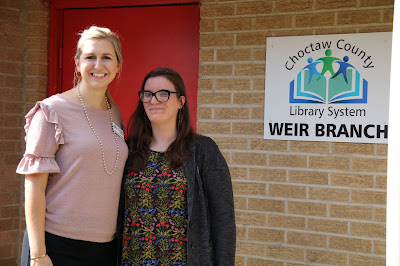 Two women stand next to each other. On the wall behind them, a sign reads, Choctaw County Library System, Weir Branch.