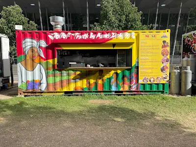 Corrogated steel storage container with a colorful mural of a woman in a headscarf on a green farm under a red and yellow sky with clouds. In the center of the structure is an open window showing a stainless steel kitchen with shelves and counters inside.