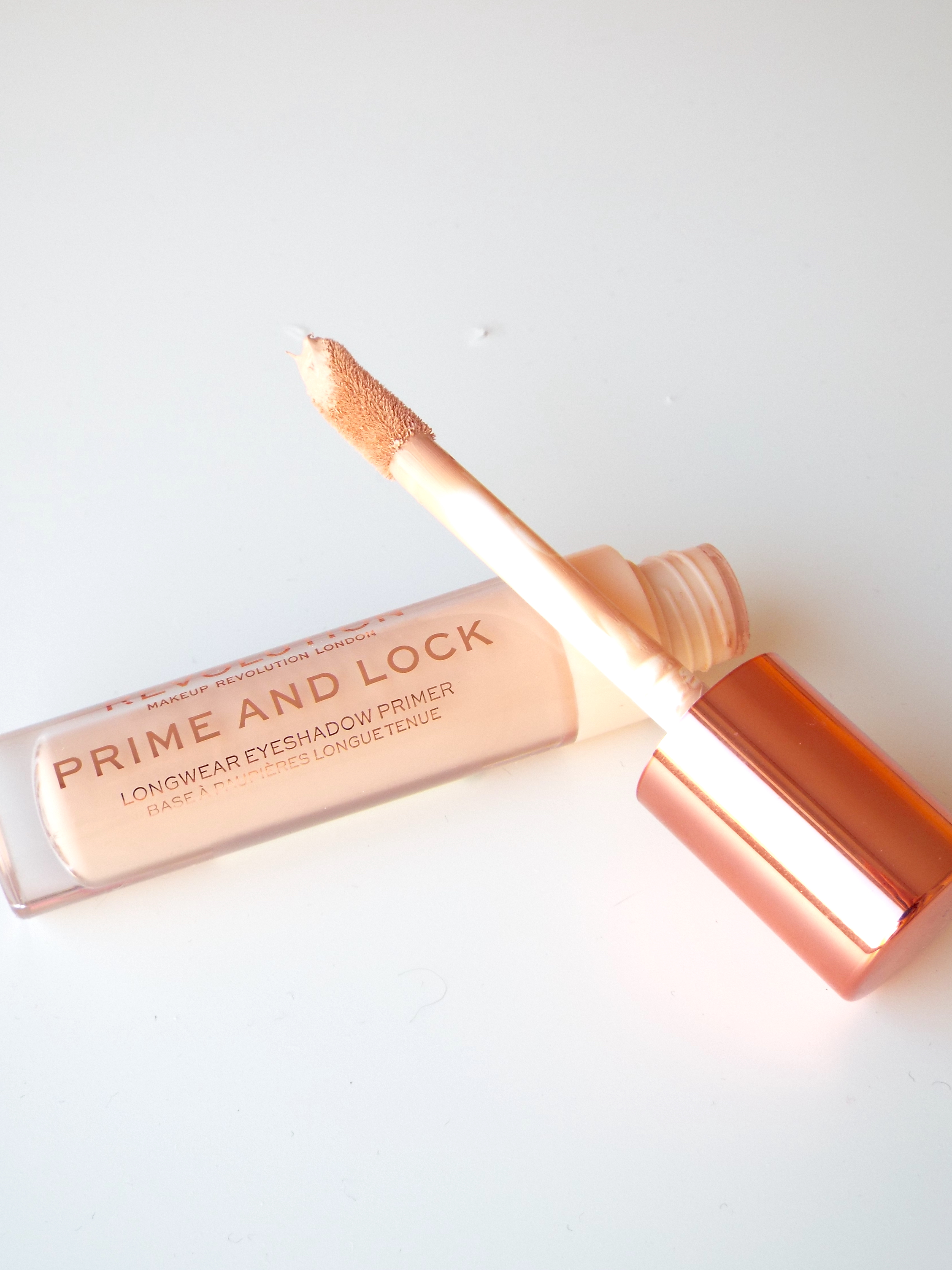 The product-coated doe foot applicator of the Makeup Revolution Prime & Lock Longwear Eyeshadow Primer, crossed over its clear plastic, rose gold bottle.