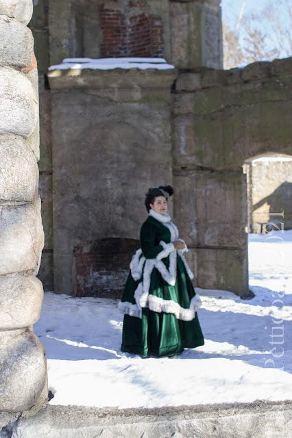 the author wearing a green velvet 170s bustle with fur trim and a gray hat in a ruined stone tower