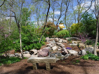 a stone bench sits at the bottom of a small hill with bushes and flowers behind it and a sculpture atop the hill