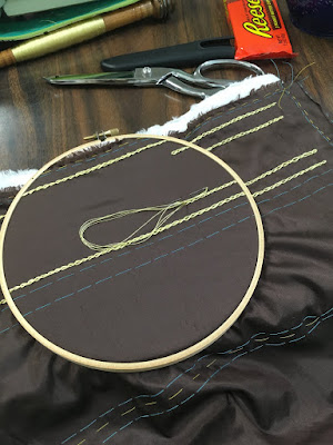 A piece of dark brown silk in a wooden embroidery hoop, with turquoise and yellow basted guidelines, two parallel rows of gold chain stitch, and two partial rows just inside the first pair. A packet of Reeses, a large pair of sewing shears, and a long wooden spool of gold thread are scattered on the table around the embroidery hoop.