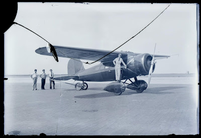 Three men stand to the left of Earhart standing on wheel of aircraft