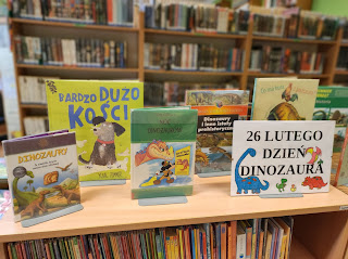 Wystawka książek o dinozaurach. Książki stoją obok siebie na półce. Tło: regały z książkami. Napis: 26 lutego Dzień Dinozaura.