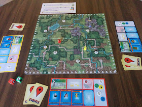 A game of Geoquest in progress. The board is in the centre, with eight cache tokens on various points, and three players' pawns are moving about the board. Each player has a player mat with point tokens, equipment cards, event cards, and travel bug tokens. The special dice sit nearby, as does a player aid.