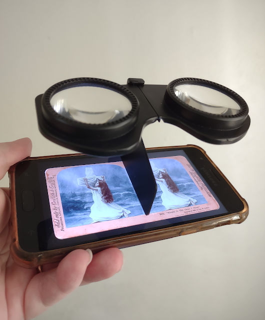 A smartphone displays a colored stereograph of a young girl clinging to a white-draped cross. Attached to the phone is a minimalist black plastic viewer consisting of a pair of lenses and a plastic clip.