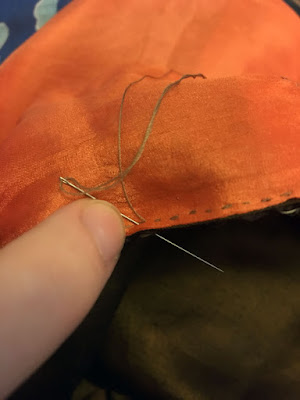 A close-up of a whit finger pushing a needle threaded with tan thread through the very edge of stacked sheer peach and green-gold fabrics, making a tiny running stitch seam.