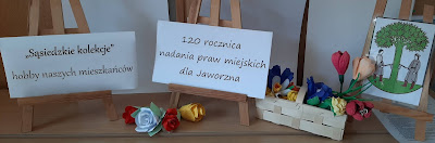 Widać 3 napisy. Pierwszy to: na białym tle czarny napis Sąsiedzkie kolekcje - hobby naszych mieszkańców. Drugi napis: na białym tle czarny napis 120 rocznica nadania praw miejskich dla Jaworzna. Trzecia kartka zawiera herb Jaworzna; na białym tle zielone drzewo i trawa a po obu stronach drzewa stoją dwaj drwale w szarym kostiumie. Obok napisów znajdują się kolorowe kwiaty wykonane z bibuły i papieru, część kwiatów w wiklinowym koszyczku a część leży luzem.