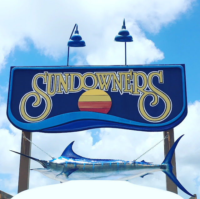 The sign for Sundowners that you see in the parking lot. It shows a color blocked sun setting into the ocean. The background of the sign is blue and large yellow letters read, "Sundowners."