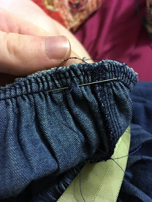 A close up of a needle inserted just below the lowest line of stitching on a machine-serged seam in blue denim, supported by a white hand.