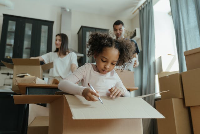 A child happily writes on a cardboard box with a pen, engaging in packing for a local move in Virginia, while her parents pack belongings in the background of their living room.