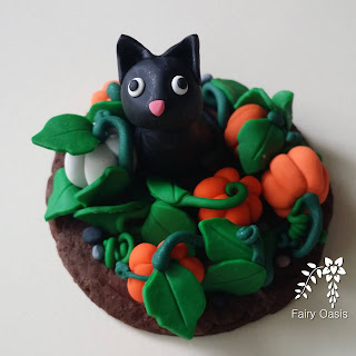[Image Description] A shimmery black cat with wide eyes sits within a patch of pumpkins gazing up either to the sky or the viewer. Everything is made of polymer clay.