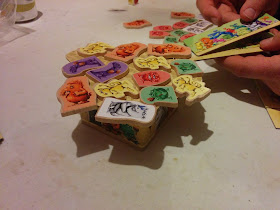 A small cardboard 'table' with an irregularly shaped tabletop. There are several wooden tokens, each adorned with a sticker of various monsters, arranged on top of this cardboard table. You can see some more of these tokens lying nearby. The hands of a player can be seen, holding the monster pusher, which consists of two rectangular pieces of cardboard, each decorated with monsters pushing at one another. The longer of the pieces is held in one hand, with a wooden monster token lying on it, and the smaller piece is used to push the wooden monster token off the bottom piece and onto the cardboard table.