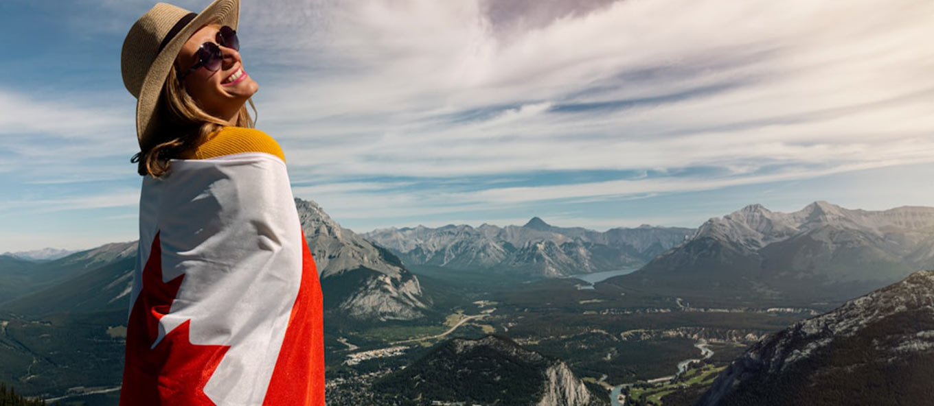 A person wearing a hat and sunglasses standing on top of a mountain in Canada with mountains in the background. The person also has a Canadian flag wrapped around them.