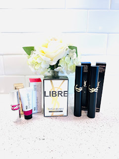 A luxurious array of YSL Beauty products arranged elegantly. From left to right: Nu Lip & Cheek Balmy Tint in a sleek, black tube; Libre Le Parfum in a sophisticated gold and black bottle; Lash Clash Extreme Volume Mascara in a bold, cylindrical container; and Rouge Volupté Candy Glaze Lip Gloss Stick in a shimmering case. The products are set against a chic, minimalist background, exuding elegance and festive charm.