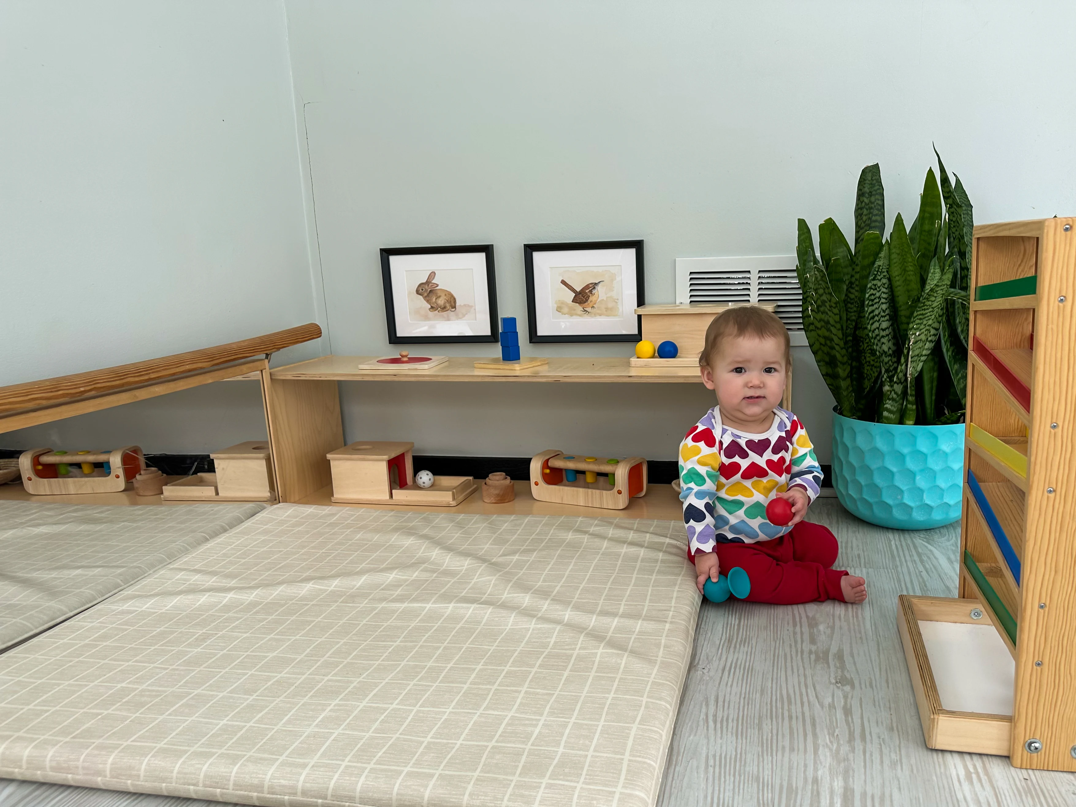 Montessori baby sits in front of playroom shelf filled with activities and toys. Montessori playspace for older baby includes mat, mirror, and wooden toys.