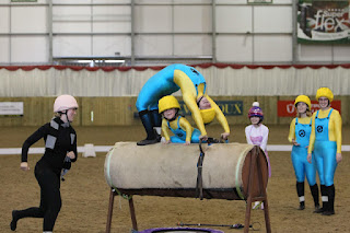 An RDA vaulting team competing on the barrel horse. Four members are dressed as minions, one is Gru and another is one of the sisters from Despicable Me. On the barrel one minion is doing a bridge and a second, smaller minion is sitting underneath the bridge in splits. The vaulter dressed as Gru is running around the side to help the smaller vaulter down. All six vaulters are grinning!