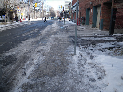 Looking along a sidewalk covered with patches of ice, where there is a significant slope down from the driveway side of the sidewalk to the road side of the sidewalk, perpendicular to the direction of pedestrian travel. (Lisgar Street just west of Elgin Street, behind the former Elgin theatre, now a coffeeshop at the corner)