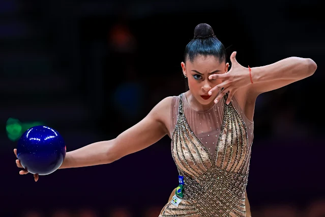 Bárbara Domingos está de collant dourado e com cara séria, segurando a bola roxa na esquerda e com a outra mão, ela tampa o lado direito do rosto. Ela é morena e negra