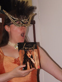 A woman in a carnival mask and orange ball gown, singing with a DVD of an opera singer