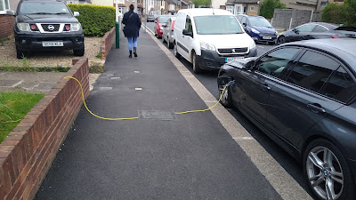 A footway with a yellow cable draped down onto it from a wall on the left and plugged into a grey car on the right. A small rubber mat covers the central section of the wire on the footway.