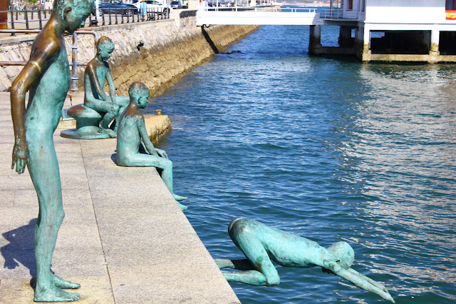 Cztery odlane z brązu figury nagich chłopców, tzw. raqueros, ustawione na nabrzeżu w Santander. Figury przedstawiające chłopców: szykujcego się do skoku, skaczącego do wody, siedzącego na brzegu, siedzącego na podporze do cumowania statków. W tle ciągnie się mur nabrzeża, widoczny jest pomost prowadzący do stojącego w wodzie na palach budynku portu jachtowego.