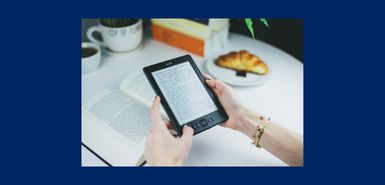 A breakfast table with a croissant and and open book and a pair of hand holing a Kindle e-reader.