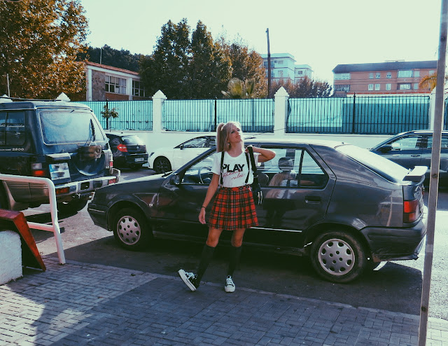 Alejandra Colomera vestida con camiseta blanca con mensaje, falda de tablas cuadros escoceses, tirantes, calcetas y zapatillas con Renault 19 de fondo