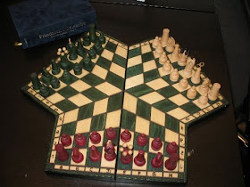 A wooden board that looks vaguely like a trefoil with squared-off leaves. Each player's section is four rows, warped into a pentagonal shape with the central columns stretching in length to reach the centre of the board, where the three sections meet. The closest section is occupied by the normal chess pieces, in red, whilst the pieces on the far right section are white and the ones on the far left are green.