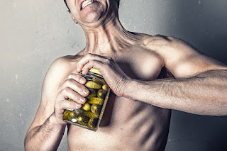 A topless white man struggles to open a jar of pickled gherkins.