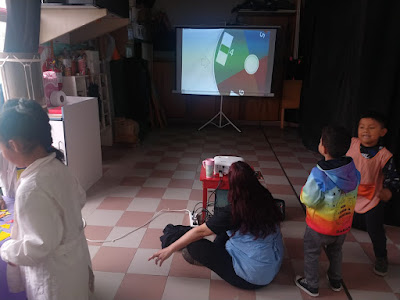 FOTO 4: MAESTRA HACIENDO GIRAR LA RULETA DIGITAL MIENTRAS UN GRUPO DE ALUMNOS JUEGA ELIGIENDO LAS FIGURAS GEOMETRICAS DEL RESULTADO DE LA MESA