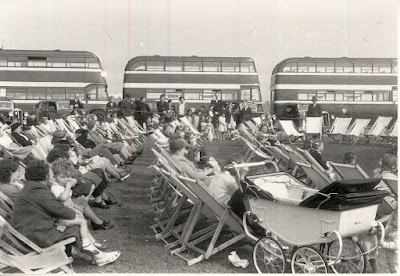 Rows of people sat in deckchairs at the seaside, with 3 double decker buses in the background