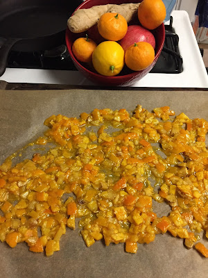 Candied lemon and orange pieces, spread out on baking paper, with a bowl of lemons, oranges, pomegranates, and ginger behind the baking sheet