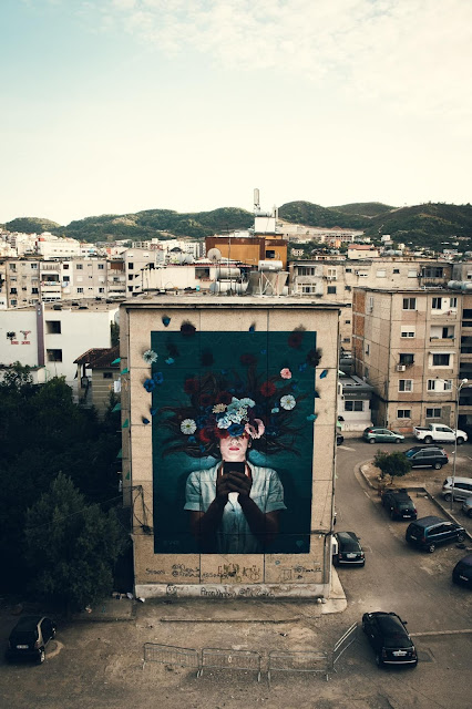 The Fabian Bane's mural in Tirana by drione where a girl is painted looking at her phone and the upper half of her face is covered with colorful flowers