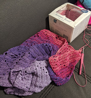 A crochet shawl work in progress scrunched up on a couch next to a box marked "Patons Summer Swirl" which has the remaining yarn in it.