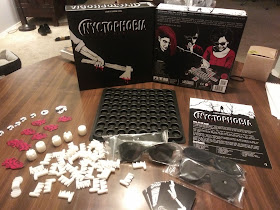 The box and components on display. The box top (the artwork represents an arm holding a bloody axe) and bottom (with artwork showing three players playing the game; two are wearing blackout glasses and the third is placing one of the other player's hand on the board) are standing behind the components: an eight by eight grid of round holes, several white plastic pieces, a small deck of cards, some cardboard tokens, the rules book, and four pairs of blackout glasses.