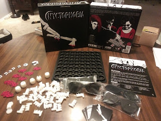 The box and components on display. The box top (the artwork represents an arm holding a bloody axe) and bottom (with artwork showing three players playing the game; two are wearing blackout glasses and the third is placing one of the other player's hand on the board) are standing behind the components: an eight by eight grid of round holes, several white plastic pieces, a small deck of cards, some cardboard tokens, the rules book, and four pairs of blackout glasses.