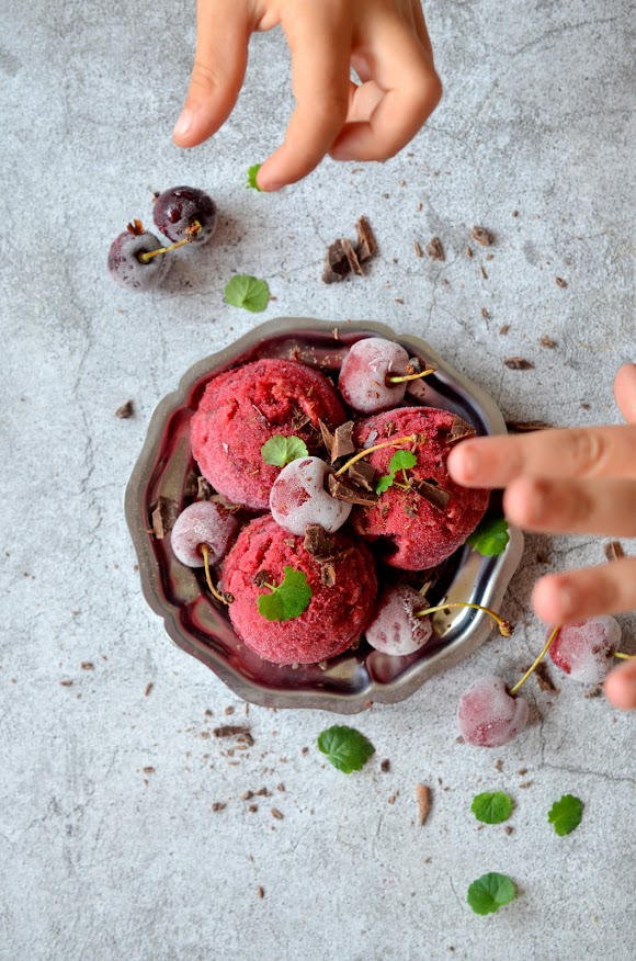 Drei Kugeln Kirschensorbet auf einem kleinen silbernen Teller, garniert mit Schokostückchen, gefrorenen Kirschen und grünen Blättchen. Zwei Kinderhände greifen nach einer Kirsche.