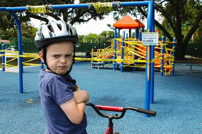 Young boy on bike wearing a helmet, with arms crossed and a scowl on his face, at the playground.